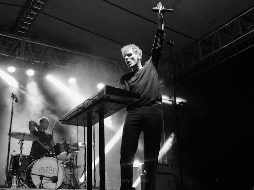 Chase Lawrence — lead singer of the band — plays the synthesizer with one hand. Ryan Winnen is shown in the background playing the drums. 