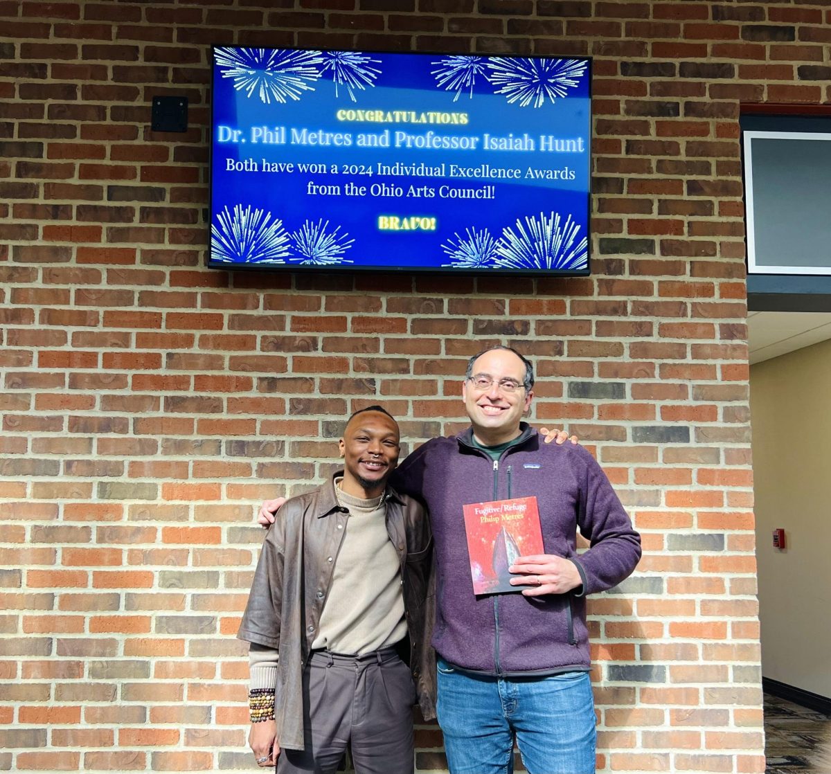 Professors Dr. Philip Metres and Isaiah Hunt celebrate their work in front of the English Department announcement TV.